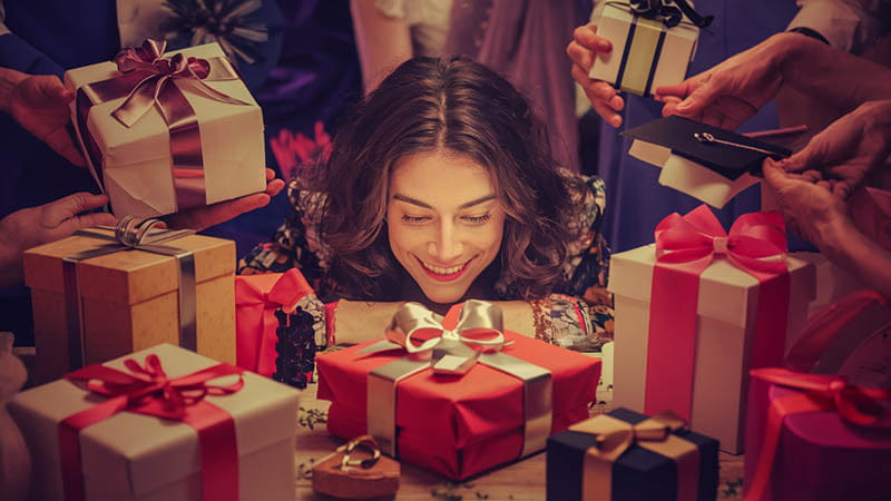 Une femme aux cheveux foncés et ondulés sourit en regardant une boîte cadeau rouge ornée d’un ruban et d’un nœud argentés dans un article intitulé comment choisir le cadeau parfait conseils simples et efficaces.