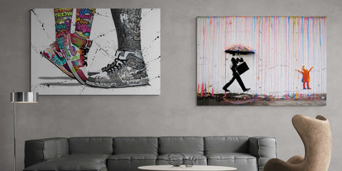 deux tableaux encadrés colorés, l’un représentant des sneakers colorées et des bottes noires avec des effets de peinture éclaboussée, l’autre une silhouette tenant un parapluie sous des coulures verticales, suspendus au mur au-dessus d’un canapé gris dans un salon.